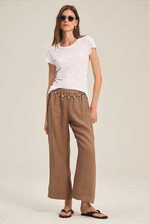 A woman in sunglasses, an ODELIA TEE by Velvet by Graham & Spencer with a classic crew neckline, brown drawstring wide-leg pants, and black flip-flops stands against a plain background—a true wardrobe essential.