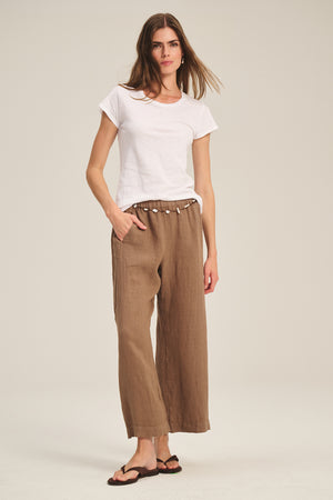 A woman stands against a neutral background wearing a white short-sleeve tee, black sandals, and the LOLA LINEN PANT by Velvet by Graham & Spencer—brown pull-on trousers with a drawstring waist crafted from lightweight linen.