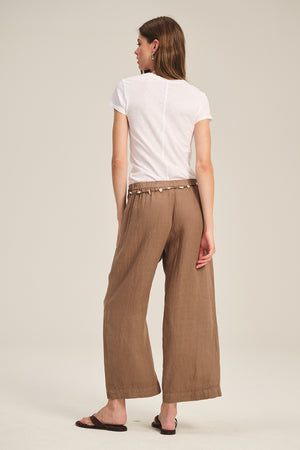 A woman stands facing away in a white t-shirt, dark sandals, and the LOLA LINEN PANT by Velvet by Graham & Spencer against a plain beige background.