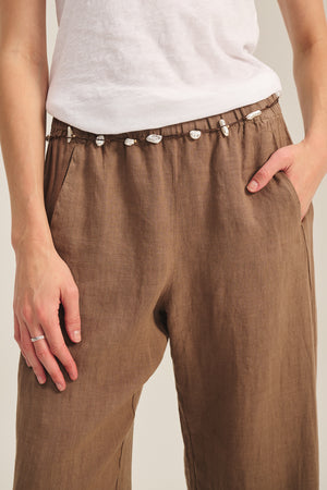 A person wearing the LOLA LINEN PANT by Velvet by Graham & Spencer, featuring a lightweight design, pull-on style, and drawstring waistband adorned with small white beads or shells, has their hands in the pockets.