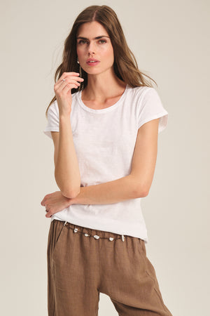 A woman poses against a neutral background wearing the ODELIA TEE by Velvet by Graham & Spencer—a cotton slub tee with a classic crew neckline—paired with brown pants, one hand near her chin and the other arm crossed.