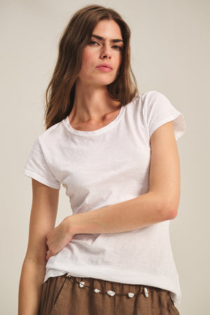 A woman with long brown hair wears the ODELIA TEE by Velvet by Graham & Spencer, featuring a classic crew neckline, paired with brown pants and standing against a neutral background.