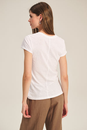 Woman stands with her back to the camera, wearing Velvet by Graham & Spencer’s ODELIA TEE—a white cotton slub crewneck—and brown pants against a plain background. A true wardrobe essential.