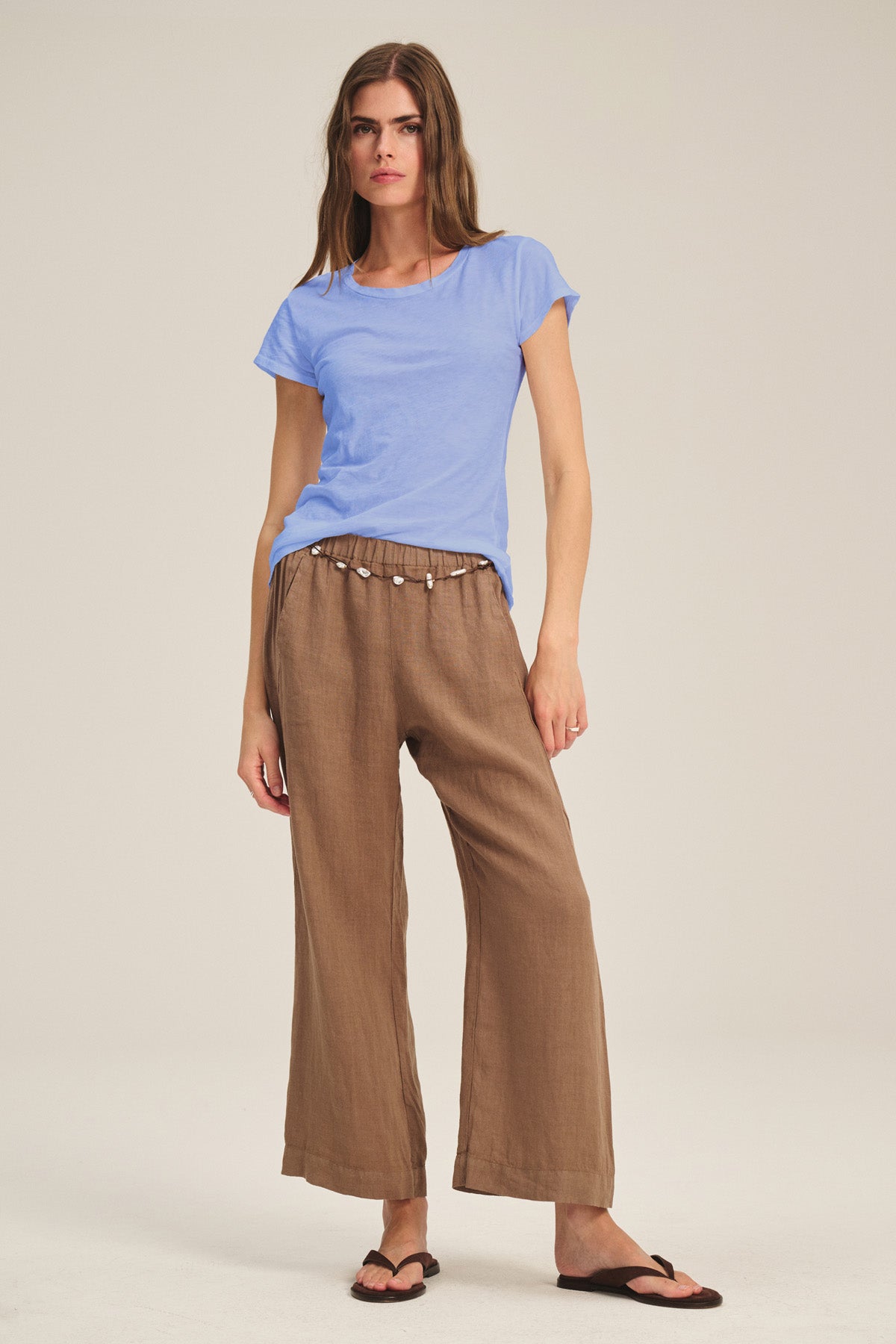 A woman stands against a plain background wearing the Velvet by Graham & Spencer ODELIA TEE in light blue, styled with brown drawstring-waist wide-leg pants and black sandals—a timeless wardrobe staple.-41015058071745