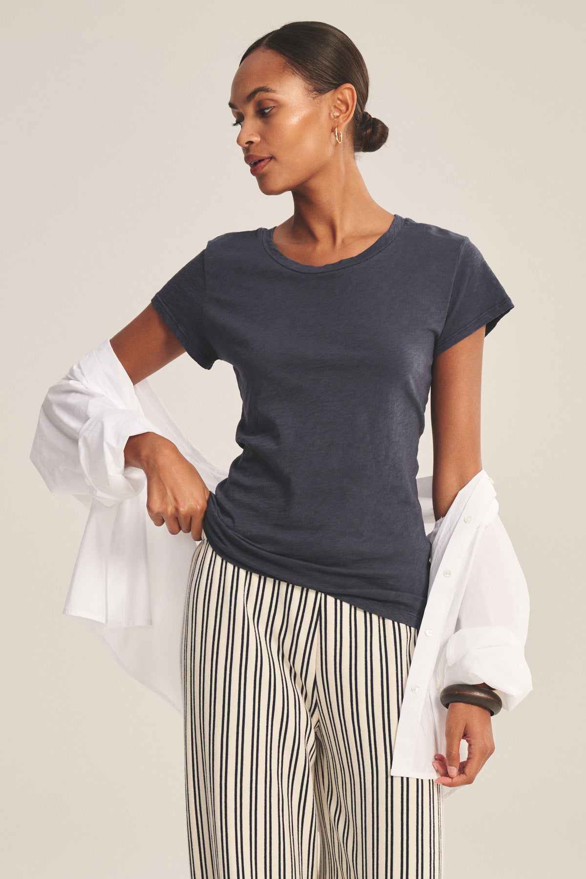 A woman stands against a neutral background in the ODELIA TEE by Velvet by Graham & Spencer—a navy cotton slub crewneck, styled with white striped pants and a white shirt draped over her arms. A true wardrobe essential.-41008381231297