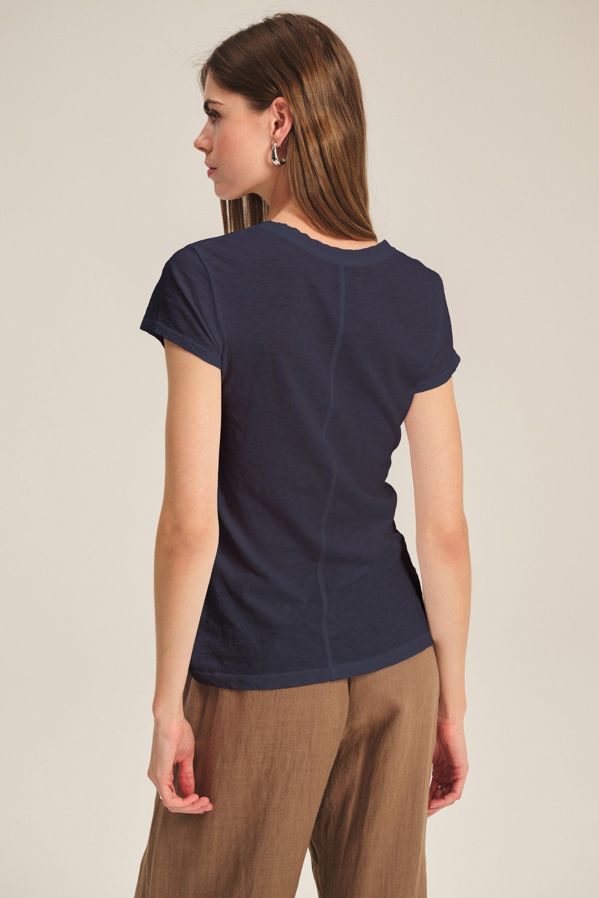 A woman faces away from the camera, wearing the ODELIA TEE by Velvet by Graham & Spencer—a navy cotton slub crew neck—paired with brown pants against a plain light backdrop.-41015058235585