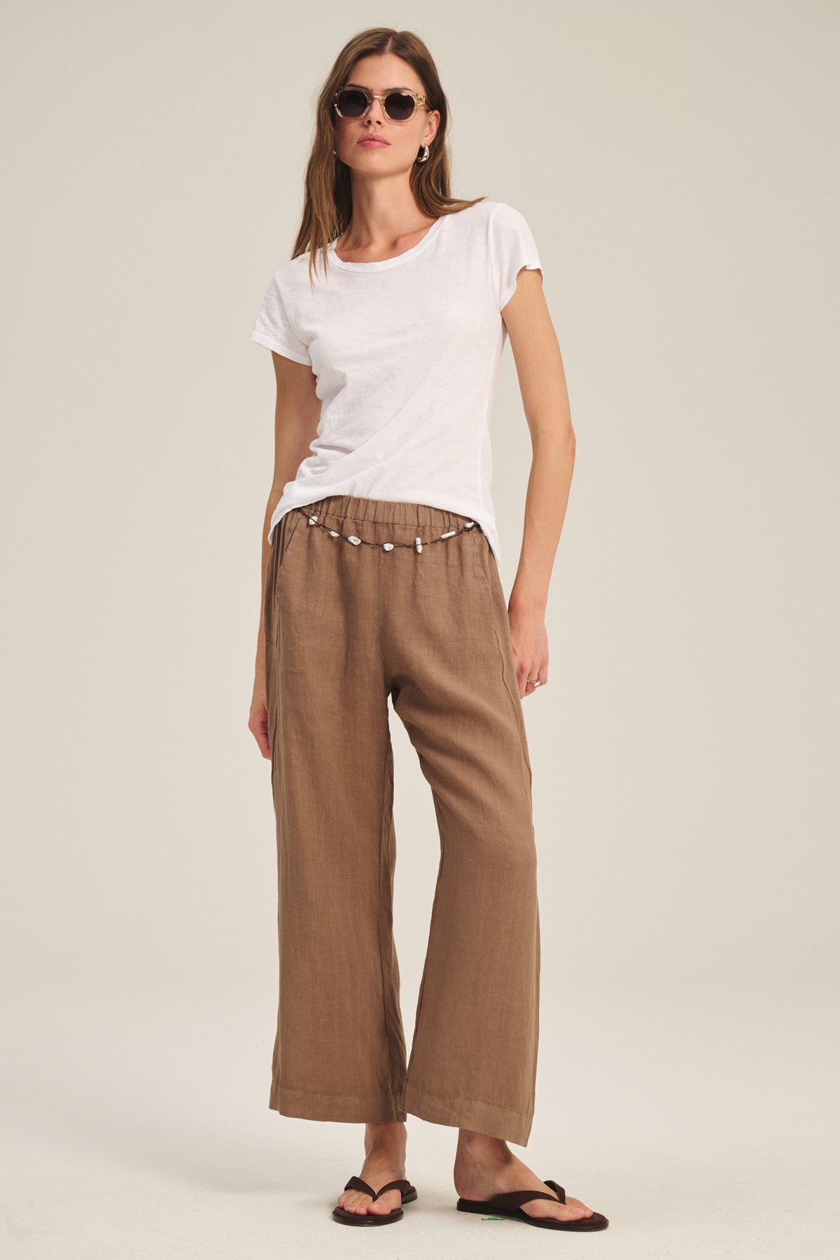 A woman in sunglasses, loose brown pants, and flip-flops wears the Velvet by Graham & Spencer ODELIA TEE—a classic crew neck white cotton slub top—standing against a plain light background.-40305622745281