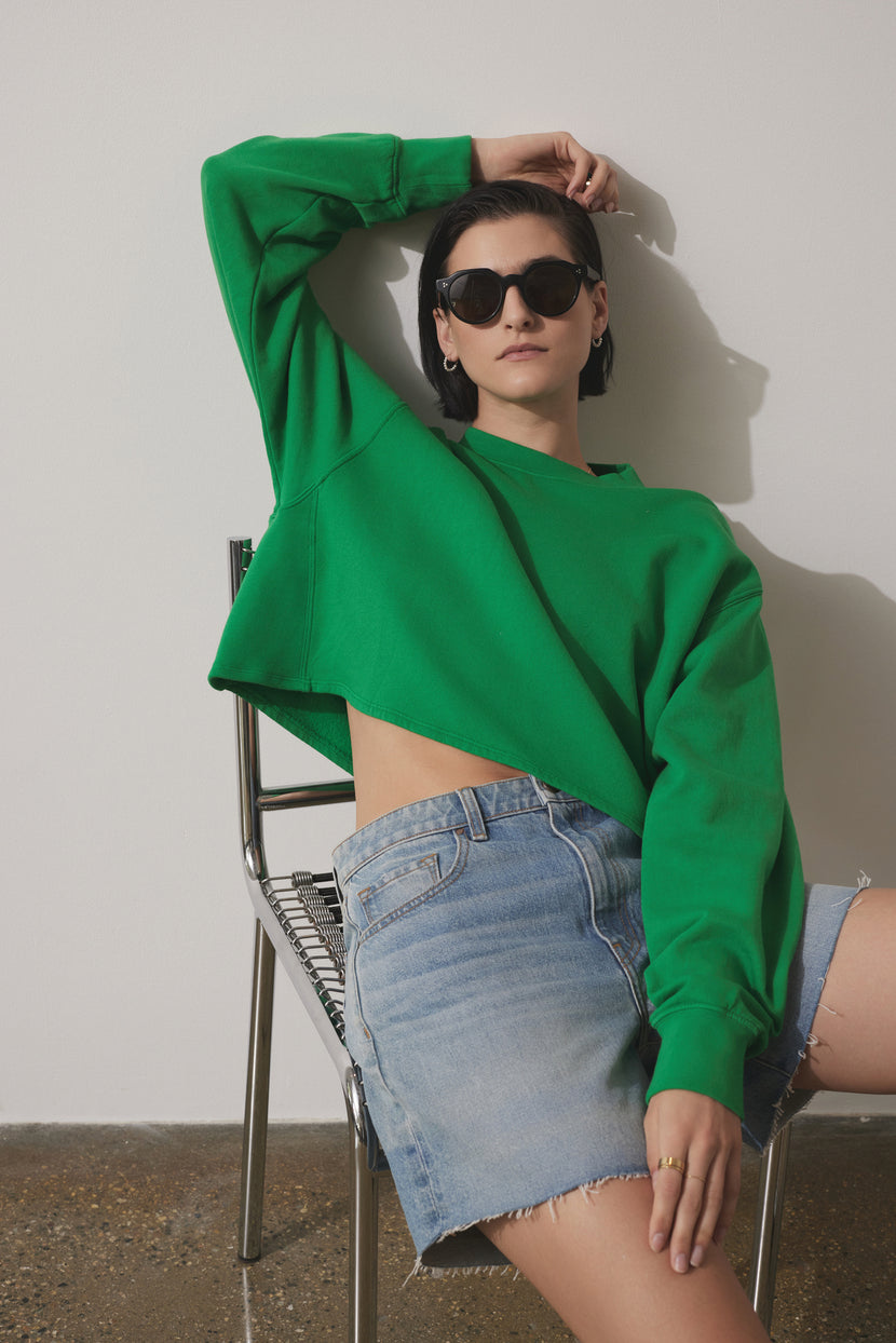 A woman wearing sunglasses, a Velvet by Jenny Graham MALIBU SWEATSHIRT with a crew neckline, and denim shorts leans back on a metal chair against a plain wall.