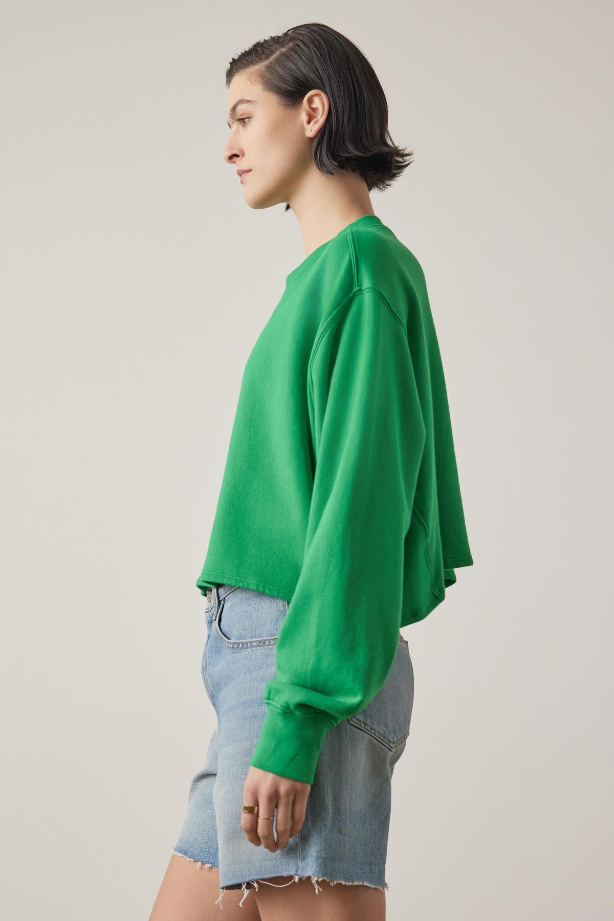   A young woman in a vibrant green Velvet by Jenny Graham Malibu sweatshirt and blue denim shorts stands in profile against a neutral background. 
