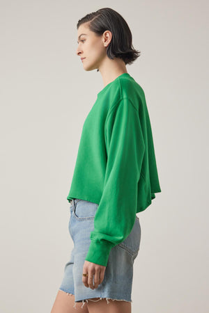 A young woman in a vibrant green Velvet by Jenny Graham Malibu sweatshirt and blue denim shorts stands in profile against a neutral background.