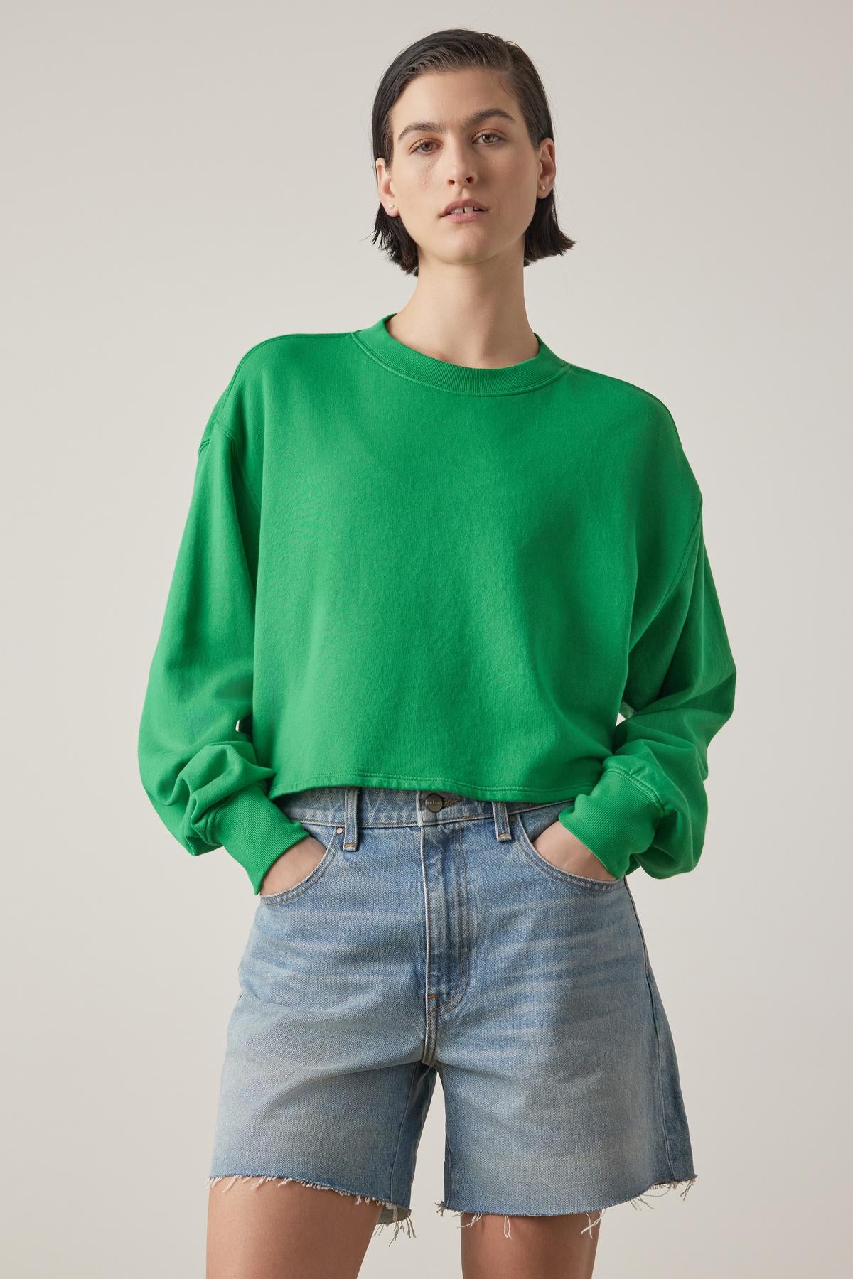   A person in a Velvet by Jenny Graham Malibu sweatshirt and blue denim shorts standing against a neutral background, hands on hips, looking at the camera. 