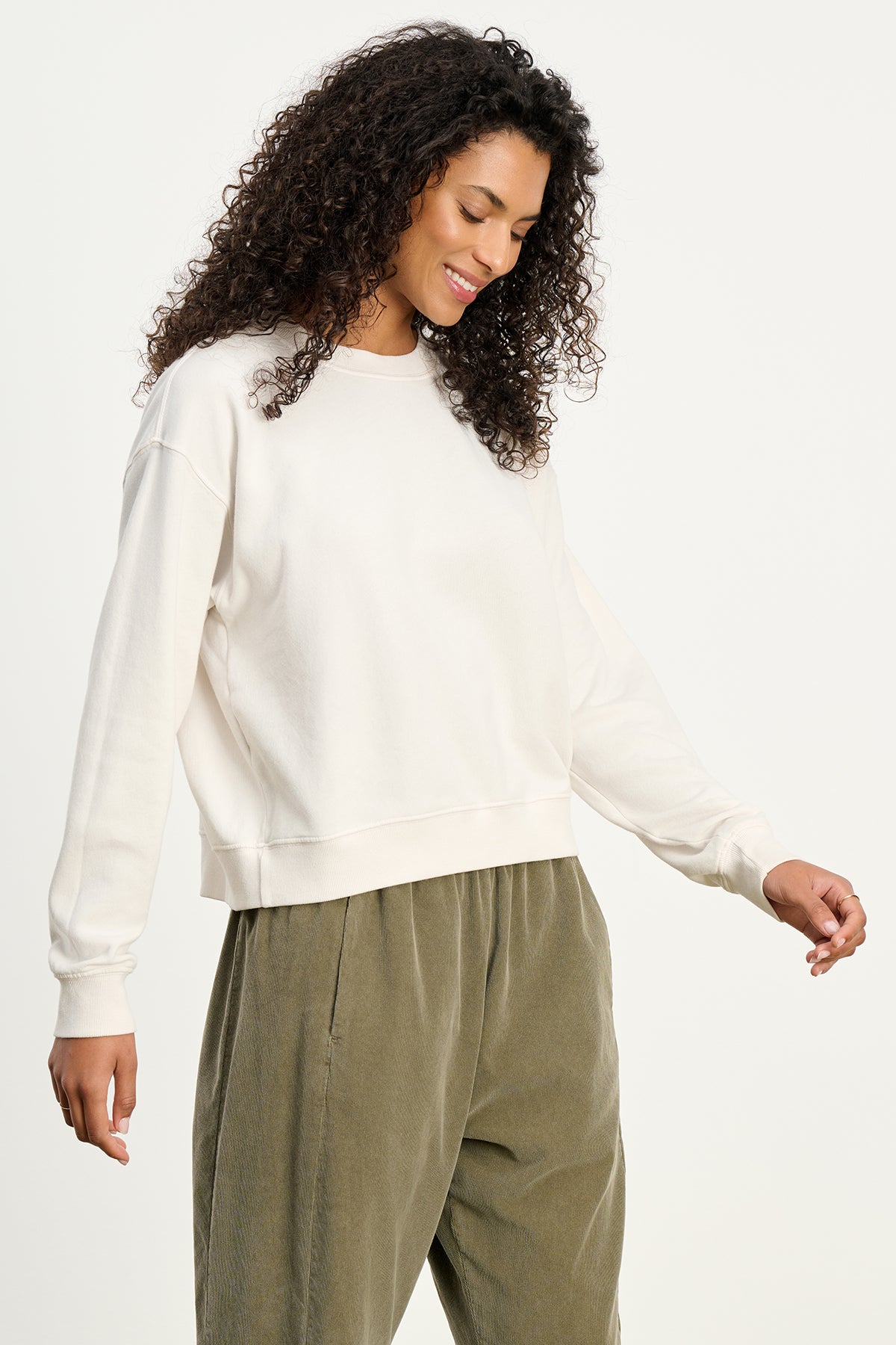 A woman with curly hair wears the YNEZ SWEATSHIRT by Velvet by Jenny Graham in off-white, paired with olive green pants, standing against a plain background and looking down with a slight smile.