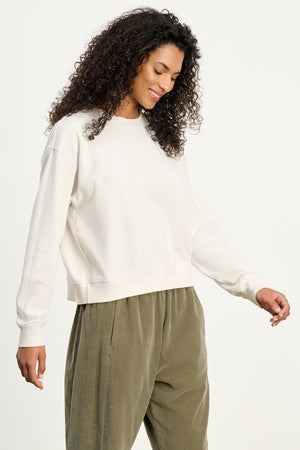 A woman with curly hair wears the YNEZ SWEATSHIRT by Velvet by Jenny Graham in off-white, paired with olive green pants, standing against a plain background and looking down with a slight smile.