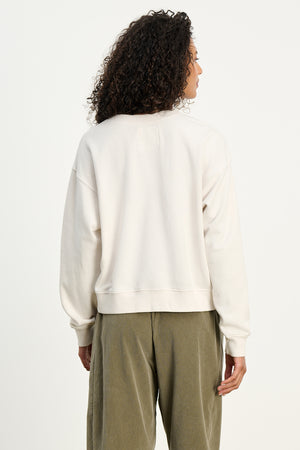 Person with curly hair wears the Velvet by Jenny Graham YNEZ SWEATSHIRT in off-white, styled with olive green pants, standing with their back to the camera against a plain background.