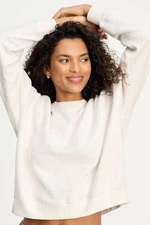 A woman with curly hair smiles and raises her arms while wearing the YNEZ SWEATSHIRT by Velvet by Jenny Graham, standing against a plain background.