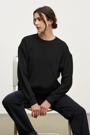 A person with dark hair in a low bun sits on a white chair, wearing the YNEZ SWEATSHIRT by Velvet by Jenny Graham and dark pants, looking to the side.