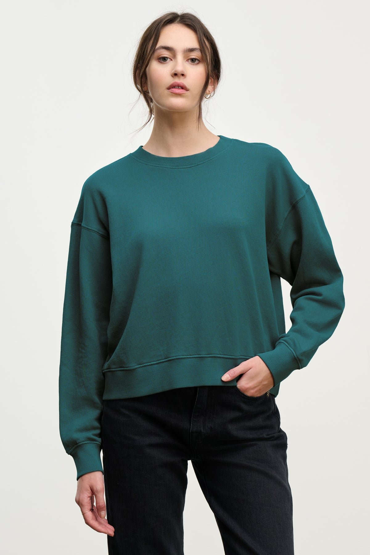 A woman wears the YNEZ SWEATSHIRT by Velvet by Jenny Graham in teal with black pants, standing against a plain light background and looking at the camera with a neutral expression.