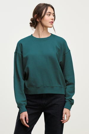Wearing the YNEZ SWEATSHIRT by Velvet by Jenny Graham in cozy teal organic cotton and black pants, a person stands against a plain light background, looking to the side.