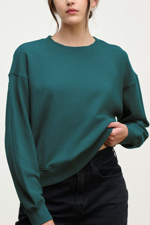 A person wearing the YNEZ SWEATSHIRT by Velvet by Jenny Graham, made from organic cotton in teal, and black jeans stands against a plain light background, holding up one side of the sweatshirt slightly.