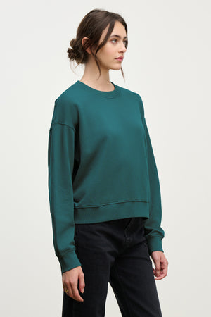 A woman with dark hair in a low bun wears the YNEZ SWEATSHIRT by Velvet by Jenny Graham, a teal cozy piece made from organic cotton, paired with black pants against a plain light background.