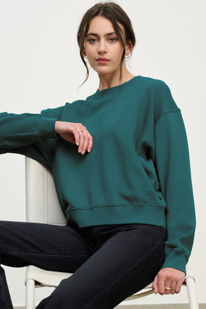 A woman with dark hair, wearing the YNEZ SWEATSHIRT by Velvet by Jenny Graham and black jeans, sits on a white chair against a plain background.