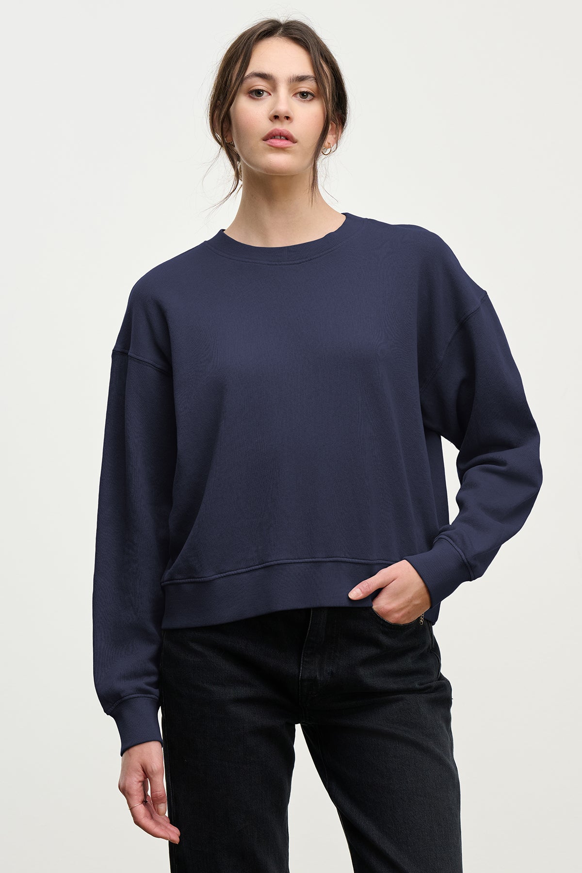 A woman in the YNEZ SWEATSHIRT by Velvet by Jenny Graham, made from organic cotton fleece, paired with black jeans, stands against a plain light background.