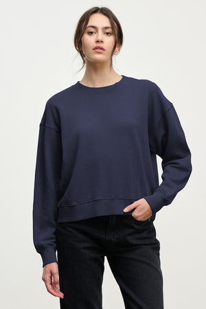 A woman in the YNEZ SWEATSHIRT by Velvet by Jenny Graham, made from organic cotton fleece, paired with black jeans, stands against a plain light background.