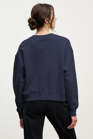 A person with brown hair in a low bun wears the Velvet by Jenny Graham YNEZ SWEATSHIRT in navy blue and black jeans, facing away from the camera against a plain background.