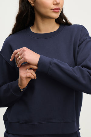 A woman wearing the Velvet by Jenny Graham YNEZ SWEATSHIRT in navy blue adjusts her sleeve cuff, with only her torso and lower face visible.