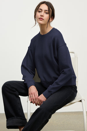 A woman sits on a white chair wearing the YNEZ SWEATSHIRT by Velvet by Jenny Graham in navy blue with black jeans, posing neutrally against a plain background.