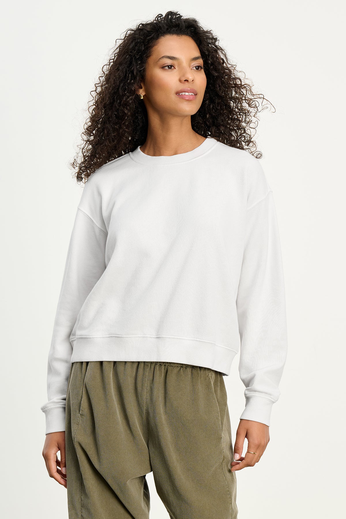 A woman with curly hair wears the YNEZ SWEATSHIRT by Velvet by Jenny Graham, a cozy white fleece cropped top, paired with loose olive green pants against a light background.