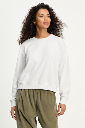 A woman with curly hair wears the YNEZ SWEATSHIRT by Velvet by Jenny Graham, a cozy white fleece cropped top, paired with loose olive green pants against a light background.