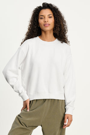 A woman with curly hair wears the cozy white YNEZ SWEATSHIRT by Velvet by Jenny Graham and olive green pants, standing against a neutral background.