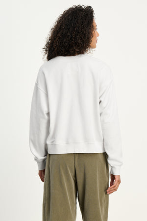 A person with curly hair is seen from behind wearing the YNEZ SWEATSHIRT by Velvet by Jenny Graham, a cozy white cropped fleece, paired with olive green pants against a plain background.