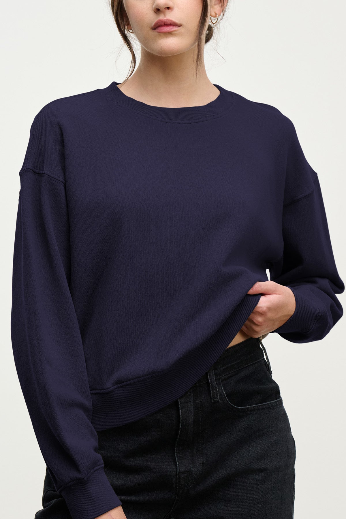 Woman wearing a loose-fitting navy blue YOKO SWEATSHIRT by Velvet by Graham & Spencer in soft cotton fleece and black jeans, standing against a plain light background, with one hand lightly pulling up the sweatshirt for a relaxed silhouette.-40431164162241