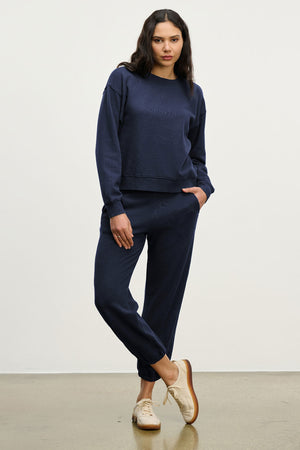 A woman poses in a relaxed stance wearing the YNEZ SWEATSHIRT by Velvet by Jenny Graham with matching jogger pants and beige sneakers against a plain white background.