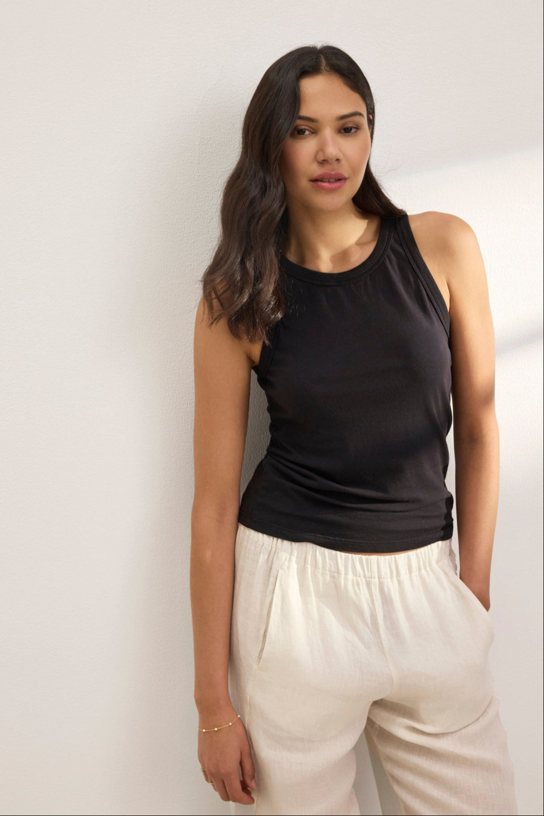 A woman with long hair in a GREER TANK TOP by Velvet by Graham & Spencer, made of organic cotton jersey, and wearing light pants leans gracefully against a white wall.-38819644965057