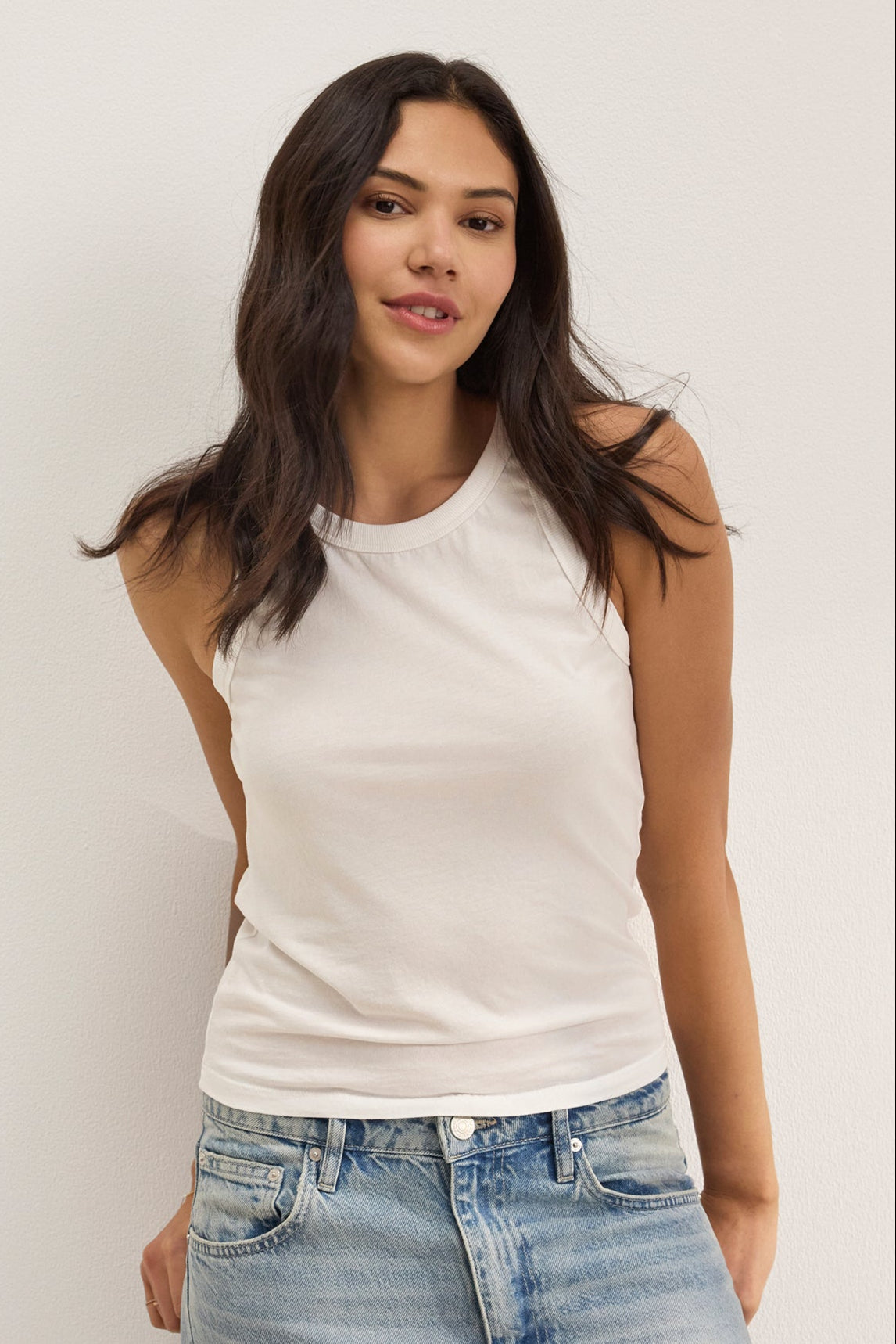 A woman with long dark hair wears the GREER TANK TOP by Velvet by Graham & Spencer, a white fitted tank with a classic crew neckline, paired with light blue jeans and standing against a plain light-colored wall.-39261747544257