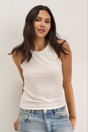 A woman with long dark hair wears the GREER TANK TOP by Velvet by Graham & Spencer, a white fitted tank with a classic crew neckline, paired with light blue jeans and standing against a plain light-colored wall.