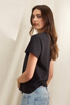 A person with long brown hair in a relaxed silhouette black JAZZ TEE by Velvet by Graham & Spencer, made from organic cotton jersey, pairs it with blue jeans while standing against a light fabric background, looking over their shoulder.