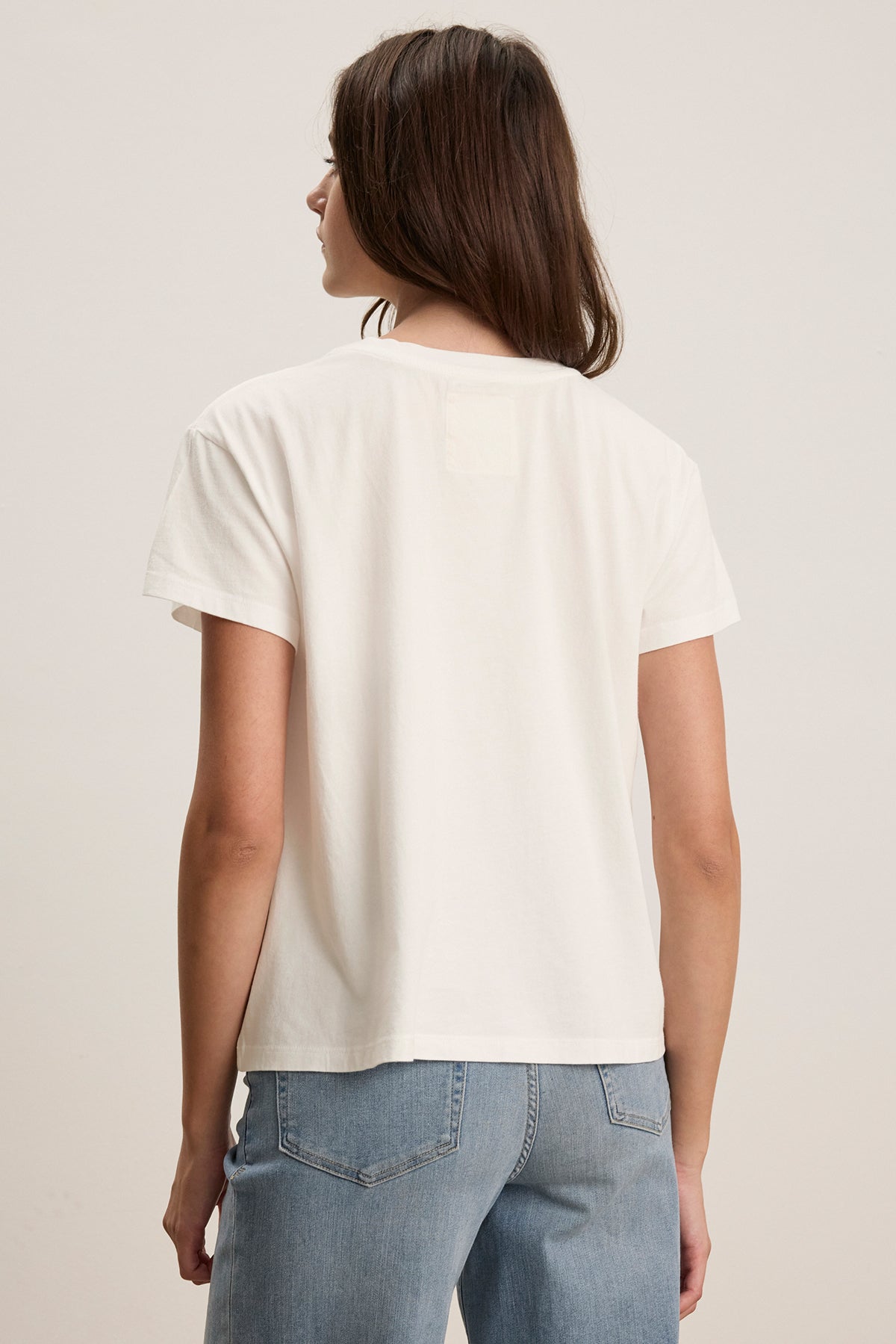 A person in a relaxed-fit TOPANGA TEE by Velvet by Jenny Graham and blue jeans faces away from the camera against a neutral background.-38866014896321