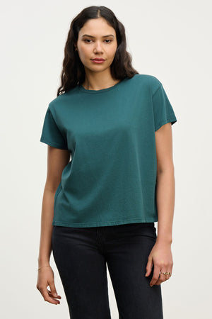 A woman with long dark hair wears the Velvet by Jenny Graham TOPANGA TEE in teal paired with black jeans, standing against a plain light background.
