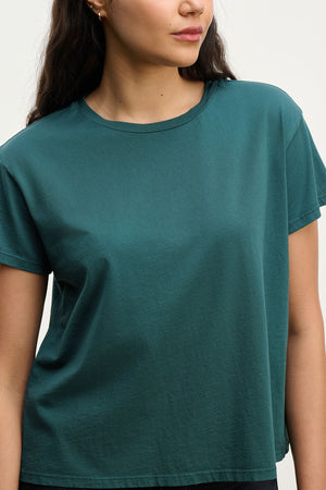 A woman stands against a plain light background wearing the TOPANGA TEE by Velvet by Jenny Graham, a relaxed-fit teal tee made from organic cotton.