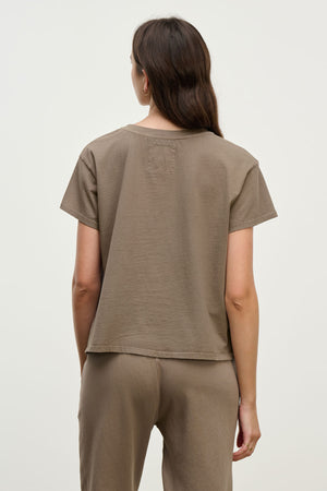 A woman faces away from the camera in the Velvet by Jenny Graham TOPANGA TEE, a relaxed-fit taupe organic cotton top—ideal for easily transitioning from work to workout against a plain backdrop.