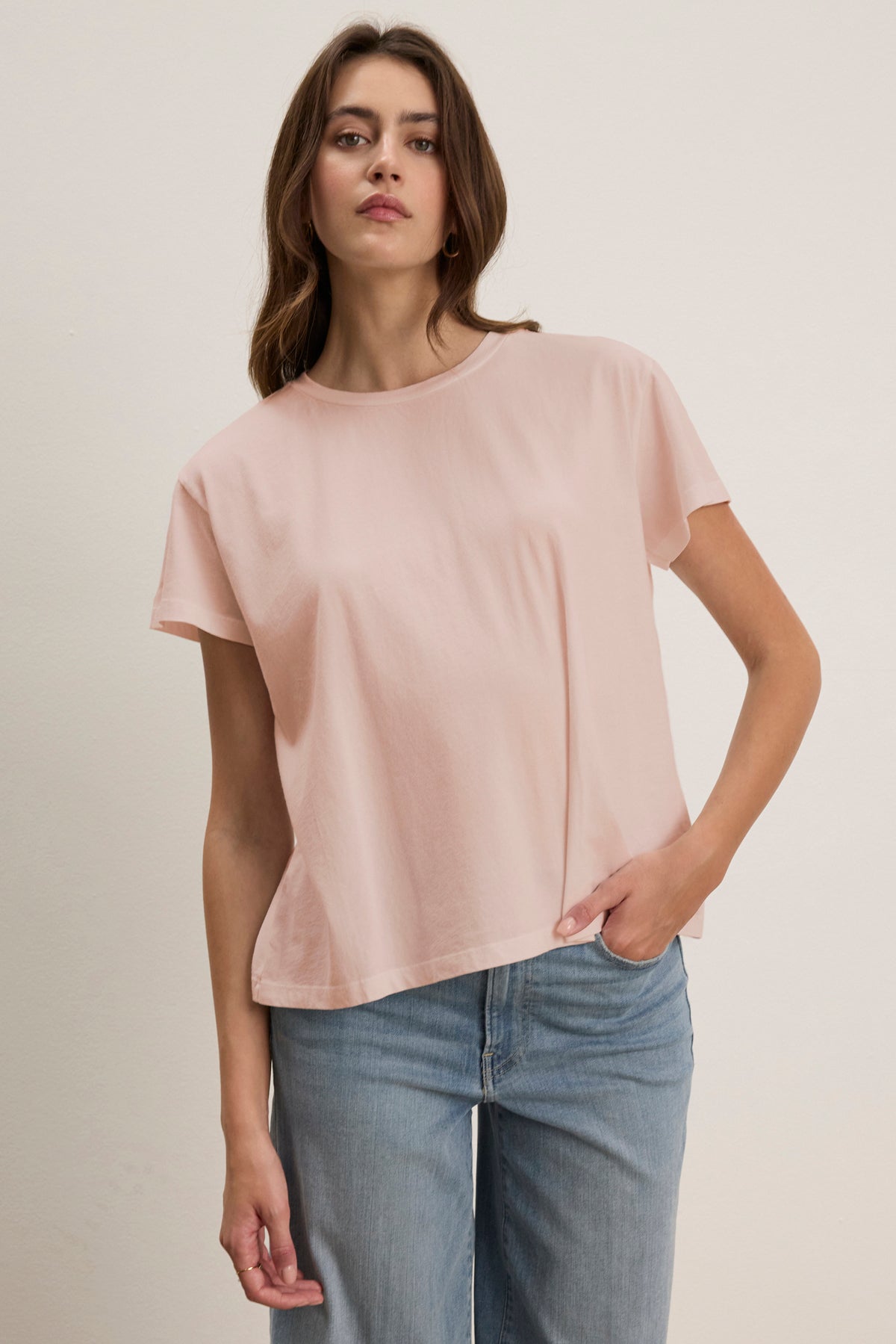 A woman stands against a white backdrop wearing the TOPANGA TEE, a light pink relaxed-fit t-shirt from Velvet by Jenny Graham, paired with jeans, proudly showcasing her affinity for sustainable clothing.-38928264462529