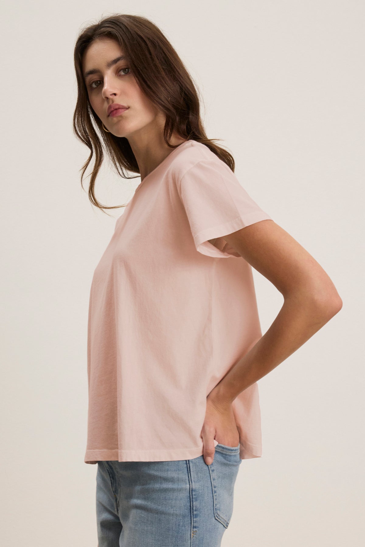 A woman poses sideways, wearing the Velvet by Jenny Graham TOPANGA TEE in light pink organic cotton and relaxed-fit blue jeans, with her left hand in her pocket against a plain backdrop.-38928264495297