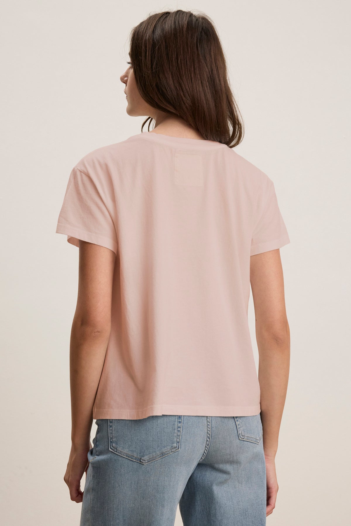 An individual in a Velvet by Jenny Graham TOPANGA TEE, light pink and made from organic cotton, with blue jeans, facing away.-38928264528065