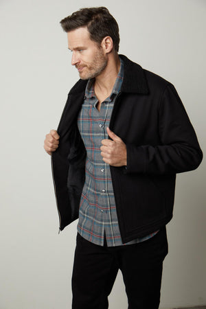 A man with short hair and facial hair wears a checkered shirt and Velvet by Graham & Spencer's CASH JACKET with a faux sherpa collar, holding the jacket open with both hands. He is looking down and to the right.