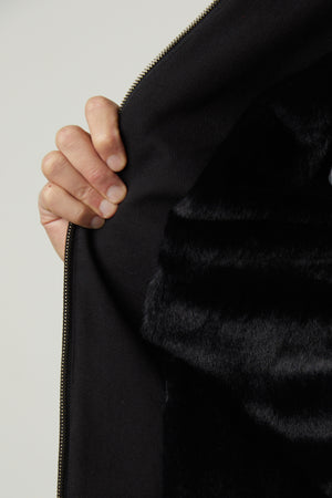 A hand holding open a black Velvet by Graham & Spencer CASH JACKET to reveal its inner black faux fur lining, complete with a warm lined interior and a sleek zipper front.