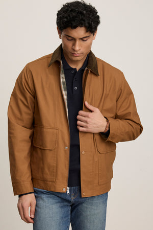 A person wears the Velvet by Graham & Spencer DESREN JACKET—brown cotton canvas with a corduroy collar—over a dark shirt and blue jeans, standing against a plain light background, looking down while adjusting the jacket.